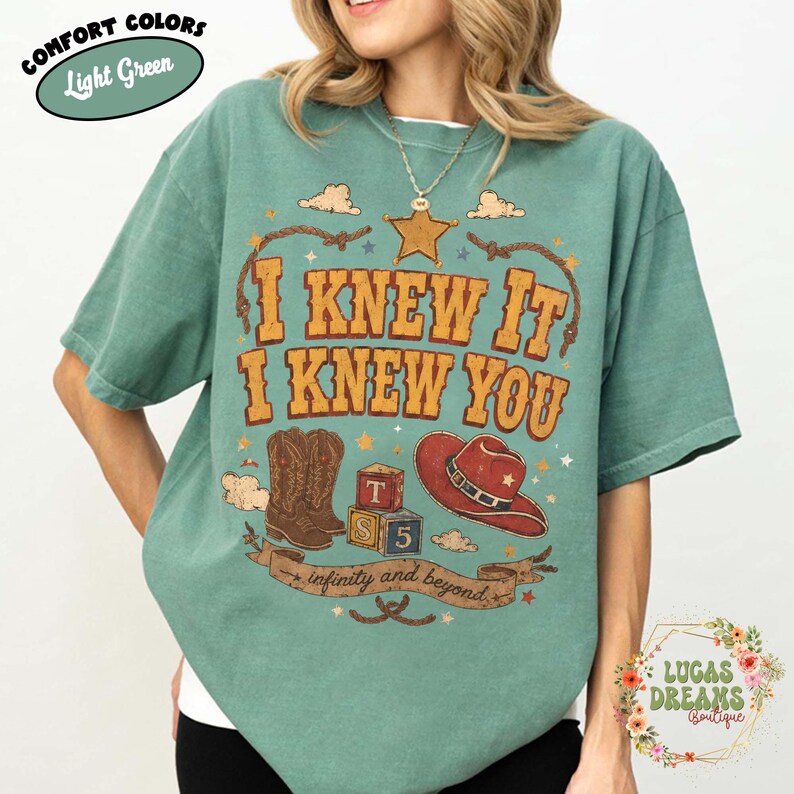 May include: A light green short-sleeve t-shirt featuring a graphic print with the text I KNEW IT I KNEW YOU and infinity and beyond. The design includes a brown cowboy boot, a red cowboy hat, a sheriff star, and two alphabet blocks labelled T and 5.