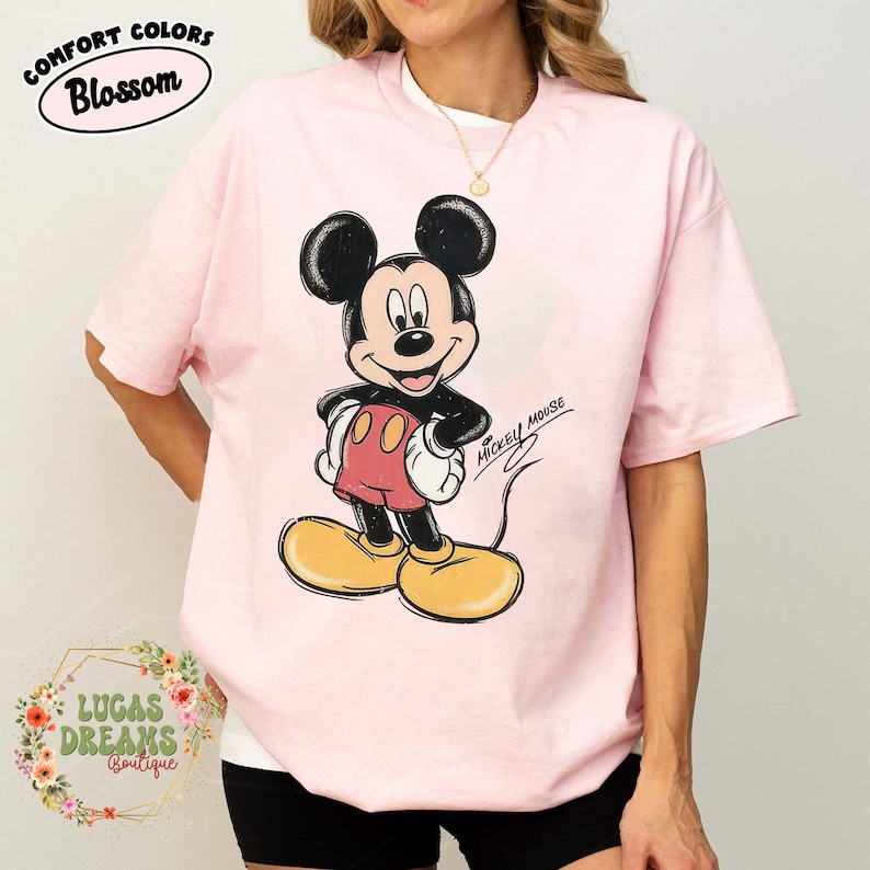 May include: A light pink short-sleeve t-shirt featuring a graphic of Mickey Mouse with black ears, red shorts, and yellow shoes. The design has a distressed texture and includes the text Mickey Mouse in a cursive script on the right side.