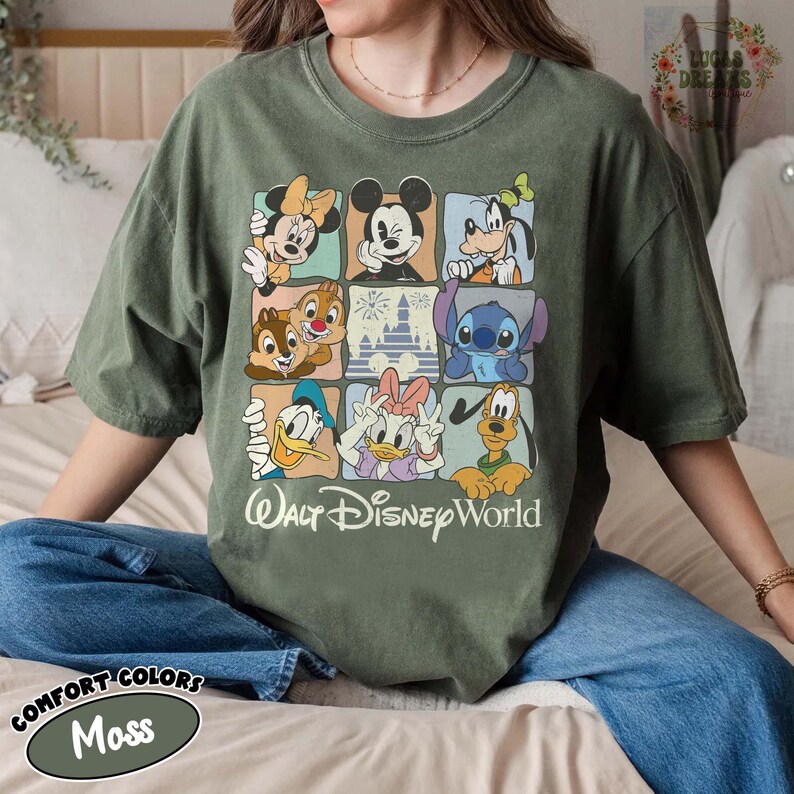 K&ouml;nnte beinhalten: Moosgr&uuml;nes T-Shirt mit einem Raster von Disney-Figuren, darunter Micky Maus, Minnie Maus, Donald Duck und Goofy. Die Worte "Walt Disney World" sind darunter gedruckt. Das Shirt ist aus Comfort Colors Material.