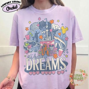 K&ouml;nnte beinhalten: Helllila T-Shirt mit einem farbenfrohen Grafikdesign, das verschiedene Freizeitpark-Symbole zeigt, darunter ein Schloss, eine Einschienenbahn und Mickey Mouse. Der Text "What Dreams Are Made Of" ist prominent. Das Shirt hat eine lockere Passform.