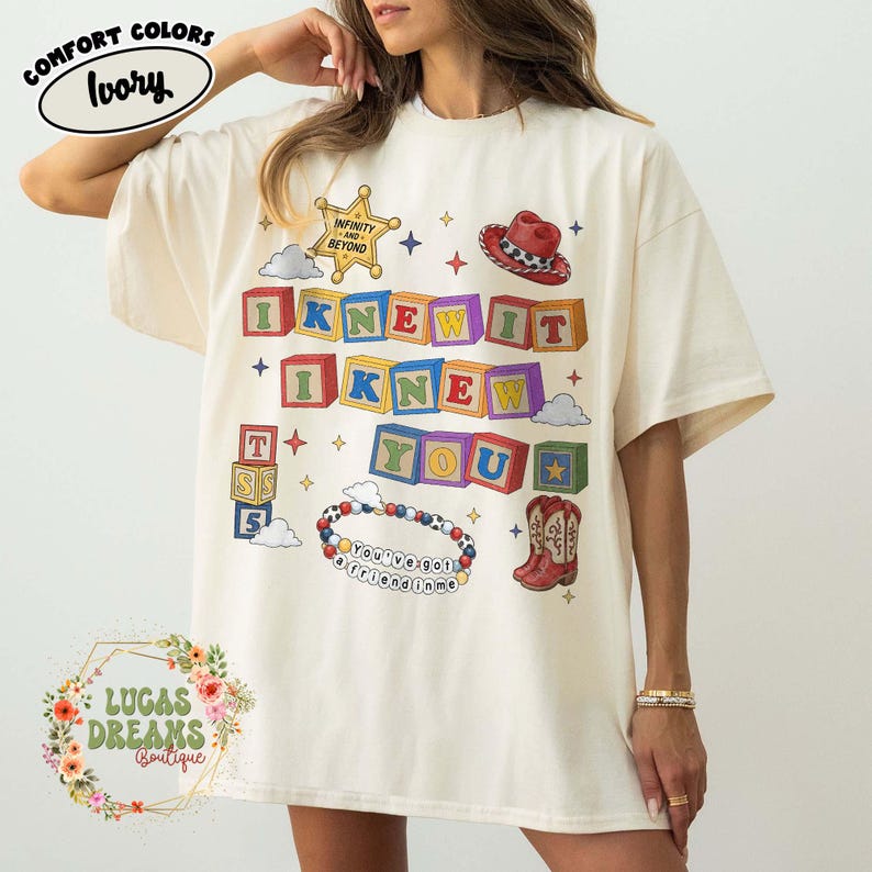 May include: An ivory short-sleeve t-shirt featuring a graphic print of colorful alphabet blocks spelling I KNEW IT I KNEW YOU. The design includes a sheriff badge with text INFINITY AND BEYOND, a cowboy hat, a beaded bracelet with text YOU'VE GOT A FRIEND IN ME, and cowboy boots.