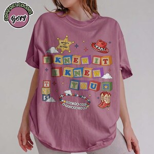 May include: A berry-colored t-shirt featuring a graphic print of colorful alphabet blocks spelling I KNEW IT I KNEW YOU. The design includes a sheriff star with the text INFINITY AND BEYOND, a cowboy hat, a pair of boots, and a beaded bracelet.