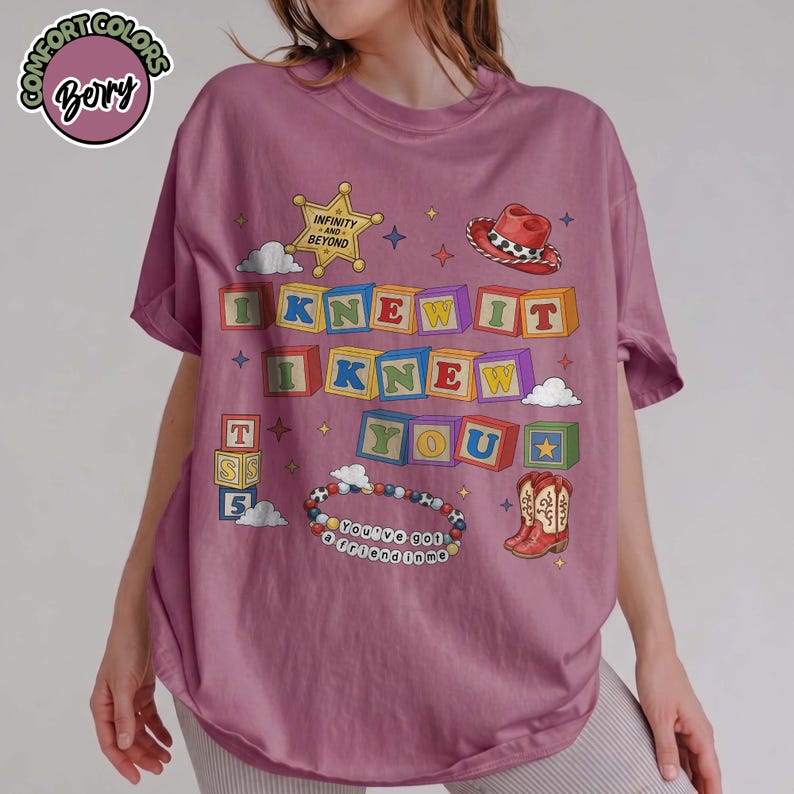 May include: A berry-colored t-shirt featuring a graphic print of colorful alphabet blocks spelling I KNEW IT I KNEW YOU. The design includes a sheriff star with the text INFINITY AND BEYOND, a cowboy hat, a pair of boots, and a beaded bracelet.