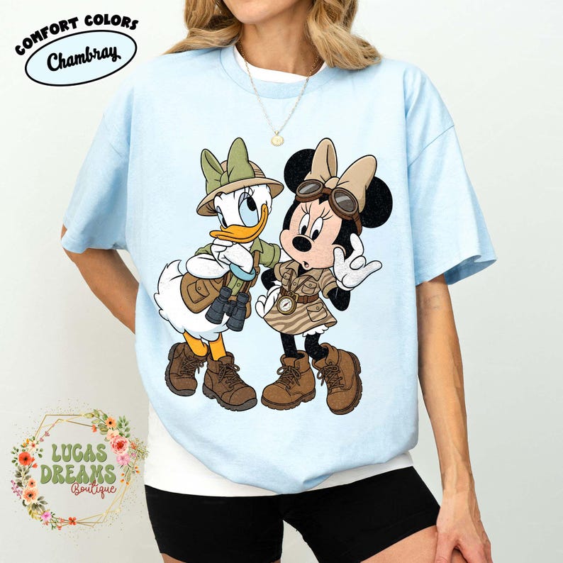 May include: Pale blue t-shirt with Daisy Duck and Minnie Mouse in safari attire. The shirt displays the text "Comfort Colors Chambray" and "Lucas Dreams Boutique". The cartoon characters are wearing safari hats, vests, and boots. The shirt is a relaxed fit.