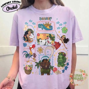 May include: A lilac t-shirt with Disney characters and the Disney logo. The shirt displays cartoon images of characters, flowers, and ocean waves. The text "Comfort Colors" and "Orchid" are at the top left.