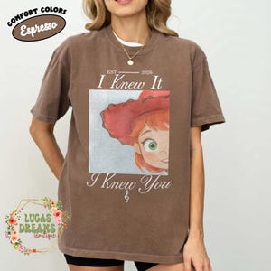 May include: An espresso brown short-sleeve t-shirt featuring a graphic print of a cartoon character with red hair and a red hat. The text reads EST 2026, I Knew It, and I Knew You, with a small musical note symbol centered below the graphic.