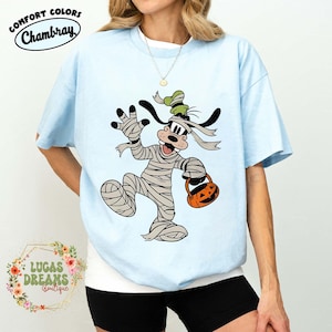 May include: Light blue Comfort Colors Chambray t-shirt featuring a Halloween-themed graphic of a cartoon dog dressed as a mummy, holding a jack-o'-lantern. The shirt also has the text "LUCAS DREAMS Boutique".