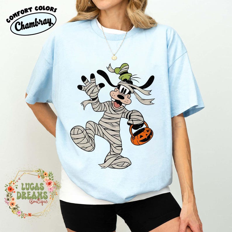 May include: Light blue Comfort Colors Chambray t-shirt featuring a Halloween-themed graphic of a cartoon dog dressed as a mummy, holding a jack-o'-lantern. The shirt also has the text "LUCAS DREAMS Boutique".