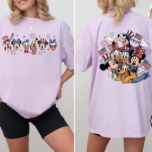 以下が含まれることがあります： 愛国的な衣装を着たディズニーキャラクターのグラフィックが特徴のラベンダー色のTシャツ。前面にはグーフィー、ミニー、ドナルド、ミッキー、デイジーが描かれています。背面には、同じキャラクターとプルートと花火が描かれています。ゆったりとしたフィット感のシャツです。