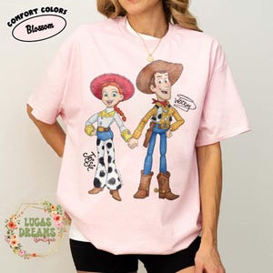 Puede incluir: Camiseta rosa claro con una ilustraci&oacute;n en acuarela de Jessie y Woody de Toy Story. Jessie lleva un sombrero rojo, una camisa blanca y pantalones de lunares blancos y negros. Woody lleva un sombrero marr&oacute;n, una camisa amarilla y pantalones azules.