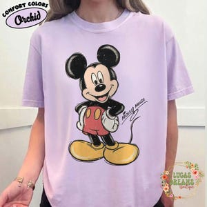 May include: A light purple short-sleeve t-shirt featuring a graphic of Mickey Mouse with black ears, red shorts, and yellow shoes. The text "MICKEY MOUSE" is printed in black script next to the character. The shirt has a crew neckline.
