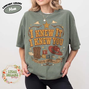 May include: A moss green t-shirt featuring a graphic print with the text I KNEW IT I KNEW YOU and infinity and beyond. The design includes a gold star, brown cowboy boots, red cowboy hat, and alphabet blocks labelled T and 5 with rope accents.