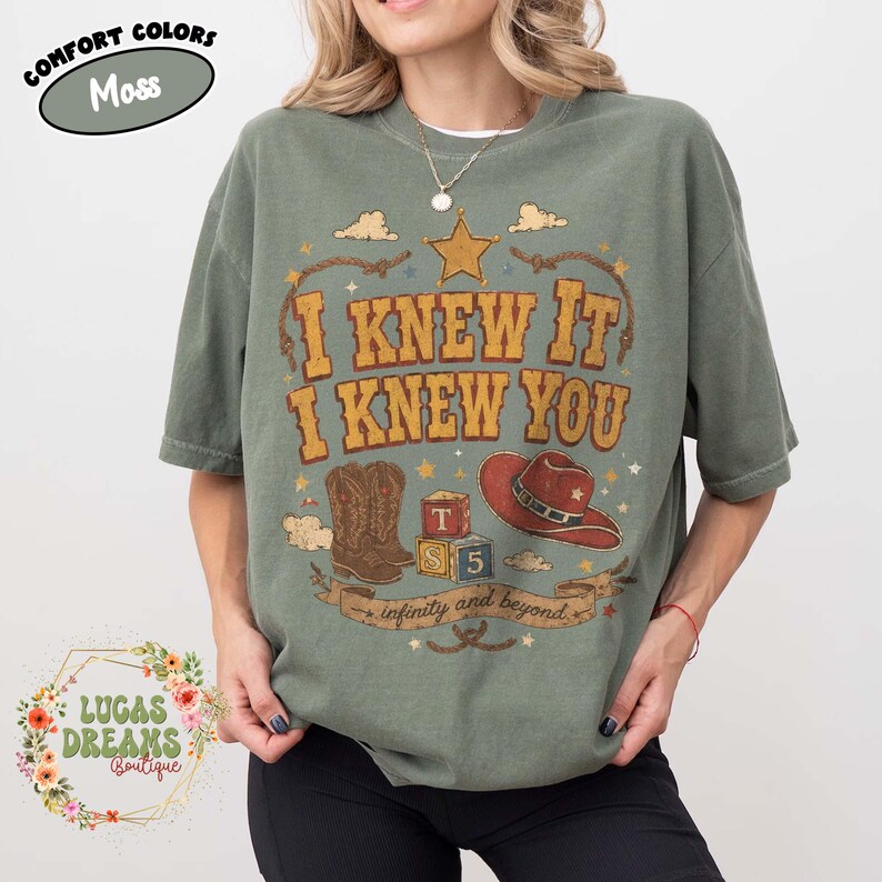 May include: A moss green t-shirt featuring a graphic print with the text I KNEW IT I KNEW YOU and infinity and beyond. The design includes a gold star, brown cowboy boots, red cowboy hat, and alphabet blocks labelled T and 5 with rope accents.