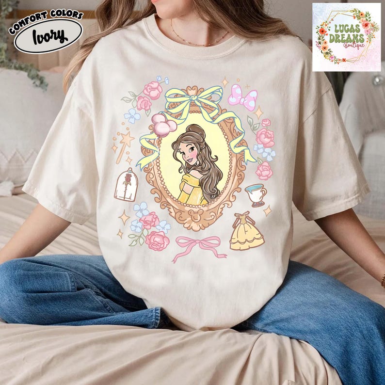 以下が含まれることがあります： アイボリー色のComfort Colors Tシャツ。黄色いドレスを着たプリンセスの漫画イラストが装飾フレームの中に描かれています。デザインにはピンクのバラ、リボン、おとぎ話の要素が含まれています。Tシャツには「Lucas Dreams Boutique」の文字が入っています。