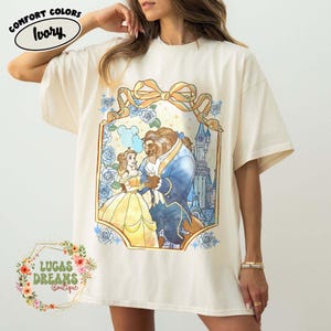 Puede incluir: Camiseta de color marfil con una ilustraci&oacute;n de estilo vidriera de Bella y la Bestia bailando. El dise&ntilde;o incluye un castillo, rosas y un gran lazo. La camiseta tiene las palabras "Comfort Colors" e "Ivory".
