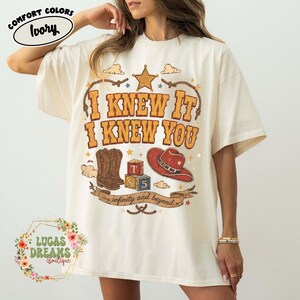 May include: An ivory t-shirt featuring a graphic print with the text I KNEW IT I KNEW YOU. The design includes a brown cowboy boot, a red cowboy hat, alphabet blocks with the letters T, S, and 5, a star, and a banner reading infinity and beyond.