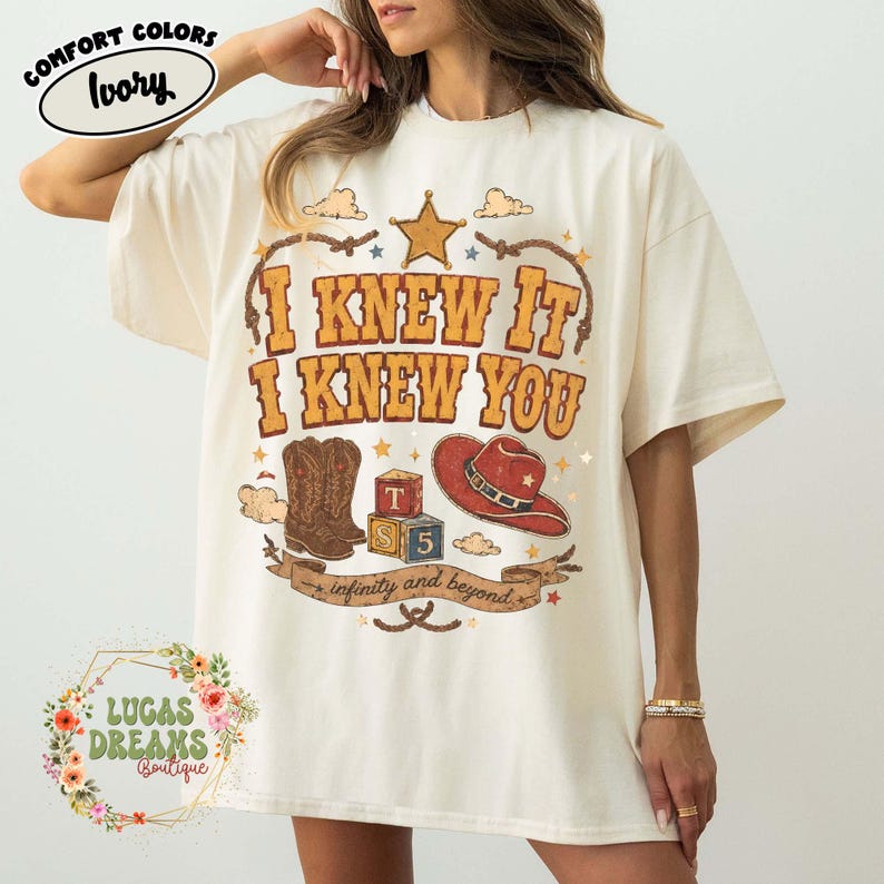May include: An ivory t-shirt featuring a graphic print with the text I KNEW IT I KNEW YOU. The design includes a brown cowboy boot, a red cowboy hat, alphabet blocks with the letters T, S, and 5, a star, and a banner reading infinity and beyond.