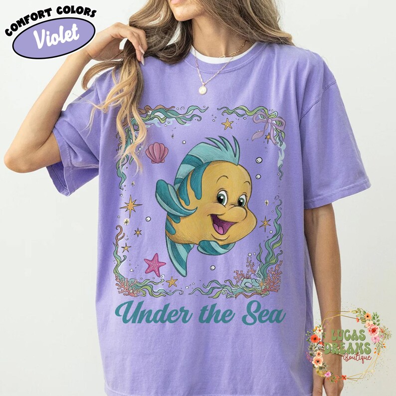 May include: Violet Comfort Colors t-shirt featuring a cartoon fish with blue and yellow details, surrounded by aquatic elements. The text "Under the Sea" is printed below the fish. The shirt has a relaxed fit.