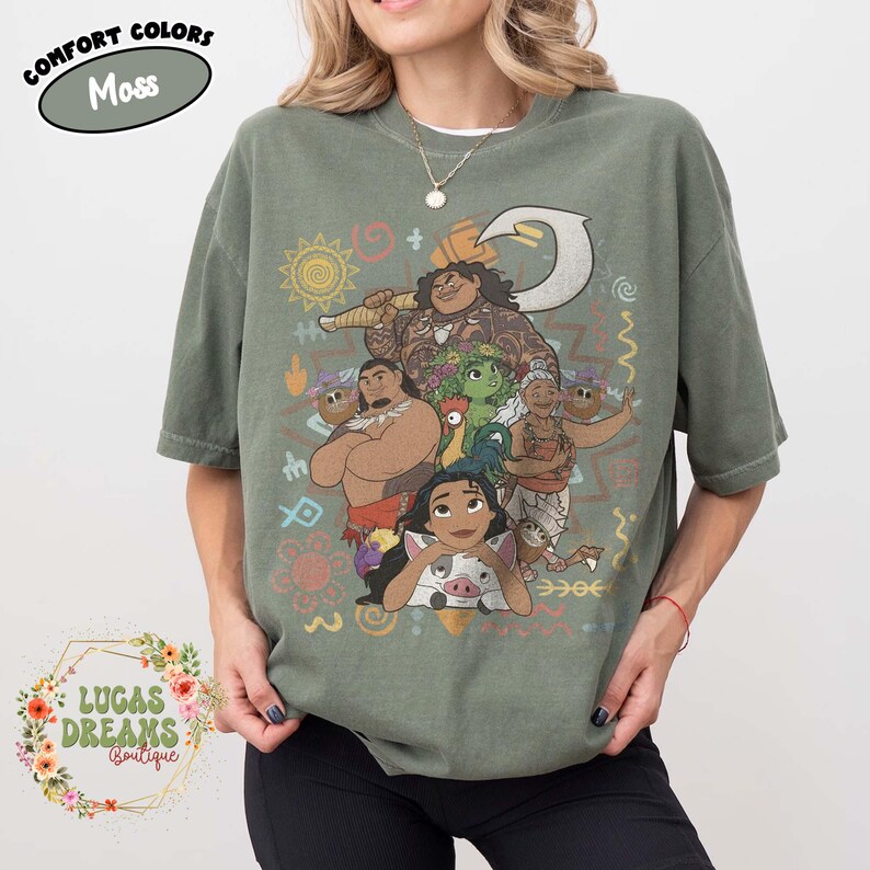 May include: A moss green short-sleeve t-shirt featuring a graphic print of characters from the movie Moana, including Moana, Maui, Pua, Hei Hei, and Gramma Tala, surrounded by various tribal-style symbols and patterns in muted earth tones.
