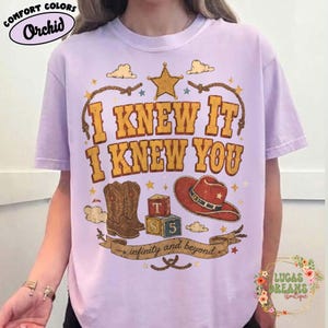 May include: A light purple short-sleeve t-shirt featuring a graphic print with the text I KNEW IT I KNEW YOU and infinity and beyond. The design includes a yellow star, a brown cowboy boot, a red cowboy hat, and two colourful alphabet blocks.