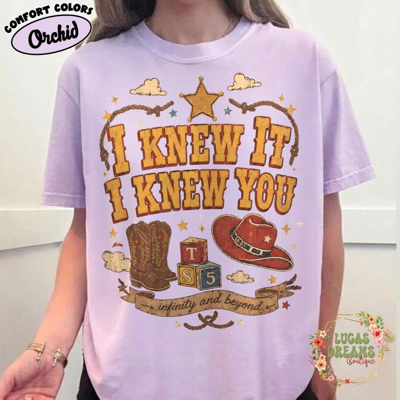May include: A light purple short-sleeve t-shirt featuring a graphic print with the text I KNEW IT I KNEW YOU and infinity and beyond. The design includes a yellow star, a brown cowboy boot, a red cowboy hat, and two colourful alphabet blocks.