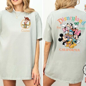 Puede incluir: Camiseta verde claro con un dibujo animado de Woody de Toy Story en la parte delantera y la palabra "Custom" debajo. La parte trasera de la camiseta muestra un gr&aacute;fico de Disneyland con Mickey, Minnie y otros personajes.