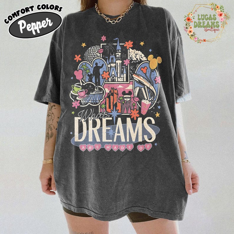 K&ouml;nnte beinhalten: Dunkelgraues T-Shirt mit einem farbenfrohen Grafikdesign mit einem Schloss, Feuerwerk und dem Text "What Dreams Are Made Of". Das Shirt hat kurze &Auml;rmel und eine lockere Passform. In der oberen linken Ecke steht der Text "Comfort Colors Pepper".