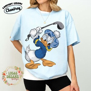 May include: Light blue t-shirt featuring a cartoon Donald Duck character swinging a golf club. The shirt has the words "Comfort Colors Chambray" at the top and "Lucas Dreams Boutique" at the bottom.