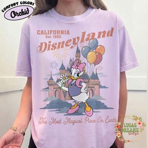 May include: Orchid-colored t-shirt with a Disneyland graphic. The design features Daisy Duck holding a flower in front of a castle, balloons, and fireworks. Text includes "California Disneyland Est. 1955" and "The Most Magical Place On Earth."