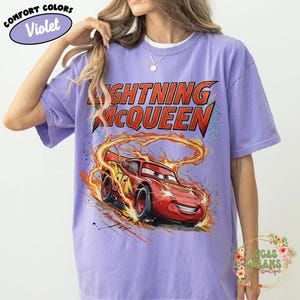 May include: Violet-colored t-shirt featuring a graphic of Lightning McQueen from the Cars movie. The design includes the character's image and the text "LIGHTNING McQUEEN" in red and yellow. The shirt has short sleeves and a crew neck.