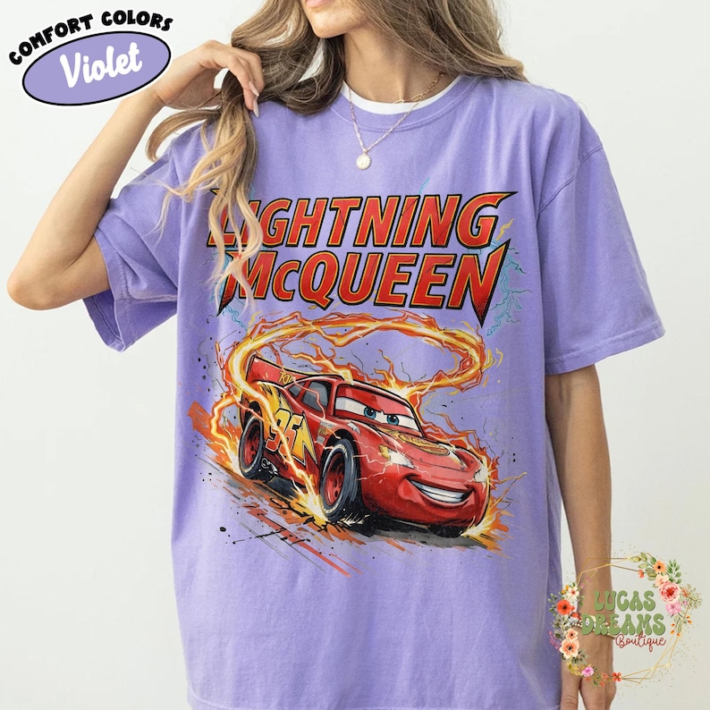 May include: Violet-colored t-shirt featuring a graphic of Lightning McQueen from the Cars movie. The design includes the character's image and the text "LIGHTNING McQUEEN" in red and yellow. The shirt has short sleeves and a crew neck.
