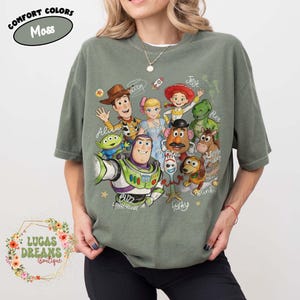 Op de afbeelding: Een mosgroen t-shirt met korte mouwen en een grafische print van diverse animatiefiguren, waaronder een cowboy, space ranger, cowgirl, dinosaurus, aardappelfiguur en hond. De tekst bevat namen zoals Woody, Jessie en Forky.