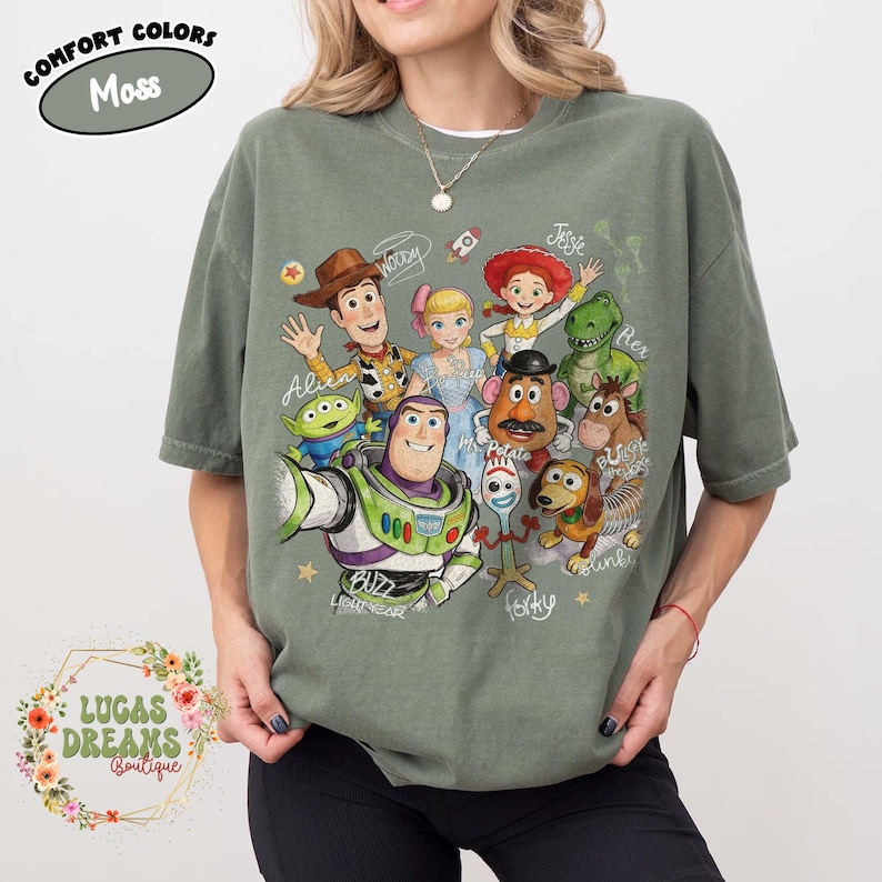 Op de afbeelding: Een mosgroen t-shirt met korte mouwen en een grafische print van diverse animatiefiguren, waaronder een cowboy, space ranger, cowgirl, dinosaurus, aardappelfiguur en hond. De tekst bevat namen zoals Woody, Jessie en Forky.