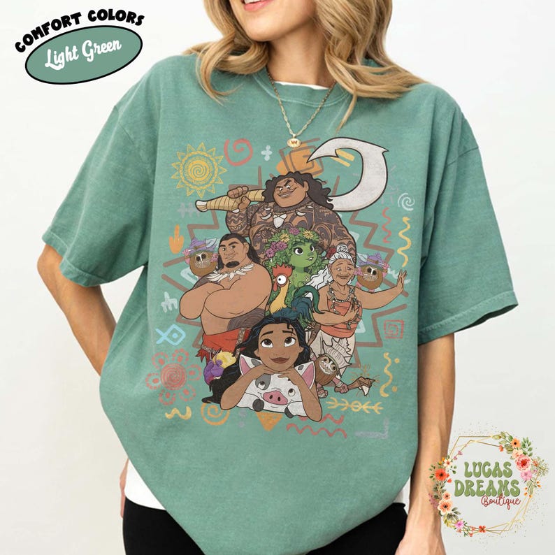 May include: A light green t-shirt featuring a graphic print of characters from the movie Moana, including Moana, Maui, Pua, Heihei, and Gramma Tala, surrounded by various tribal-style symbols and patterns in orange, yellow, and white colors.
