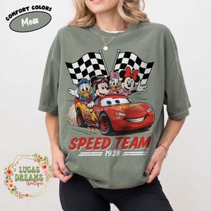 Op de afbeelding: Mosgroen t-shirt met een afbeelding van Disney-personages Mickey Mouse, Donald Duck, Daisy Duck en Minnie Mouse in een rode raceauto. De tekst "SPEED TEAM 1928" staat onder de auto gedrukt, met geblokte vlaggen op de achtergrond.