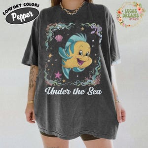 May include: Dark gray t-shirt featuring a cartoon Flounder from The Little Mermaid, surrounded by aquatic elements. The text "Under the Sea" is printed below the graphic. The shirt has a vintage, worn look.
