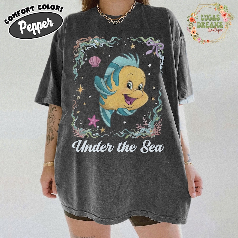 May include: Dark gray t-shirt featuring a cartoon Flounder from The Little Mermaid, surrounded by aquatic elements. The text "Under the Sea" is printed below the graphic. The shirt has a vintage, worn look.