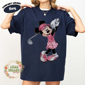 May include: Navy blue t-shirt featuring Minnie Mouse in a pink golf outfit, swinging a golf club. The shirt has the words "Comfort Colors" and "Navy" printed on it. The shirt also has the words "LUCAS DREAMS Boutique" printed on it.