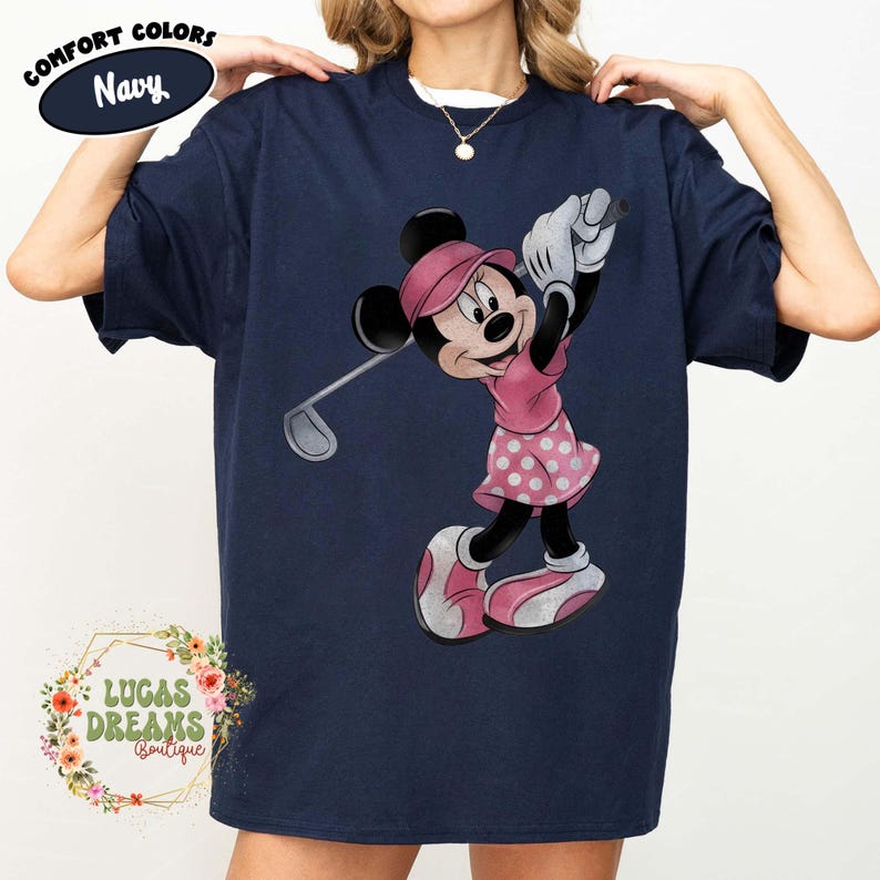 May include: Navy blue t-shirt featuring Minnie Mouse in a pink golf outfit, swinging a golf club. The shirt has the words "Comfort Colors" and "Navy" printed on it. The shirt also has the words "LUCAS DREAMS Boutique" printed on it.