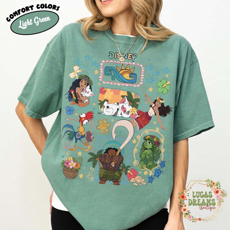 May include: Light green t-shirt featuring Disney characters. The shirt displays illustrations of Moana, Maui, Hei Hei, Pua, and other characters. The text "Disney" is printed across the chest. The shirt also has the text "Comfort Colors Light Green".