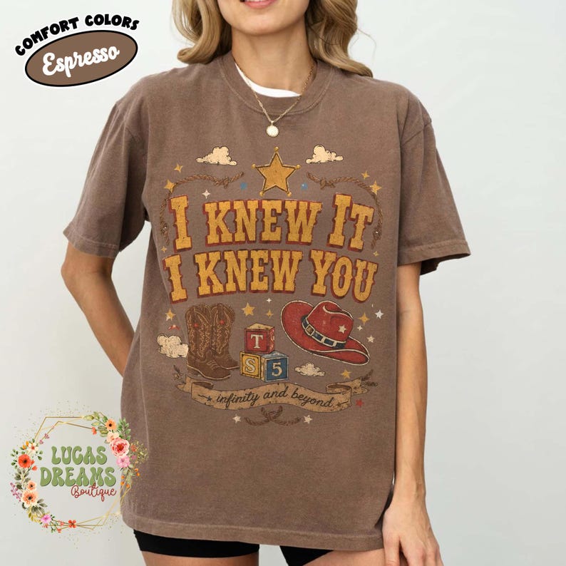 May include: An espresso brown short-sleeve t-shirt featuring a graphic print with the text I KNEW IT I KNEW YOU and infinity and beyond. The design includes a yellow star, a pair of brown cowboy boots, a red cowboy hat, and alphabet blocks.