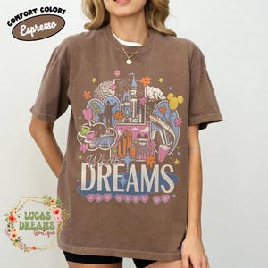 K&ouml;nnte beinhalten: Braunes T-Shirt mit einem farbenfrohen Grafikdesign mit einem Schloss, Wahrzeichen und dem Text "What Dreams Are Made Of". Das Shirt hat kurze &Auml;rmel und einen Rundhalsausschnitt. In der oberen linken Ecke steht "Comfort Colors Espresso."