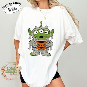 May include: White t-shirt featuring a green alien dressed as a mummy holding an orange jack-o'-lantern. The shirt has the words "COMFORT COLORS" and "White" in a circle at the top. The shirt is from "LUCAS DREAMS Boutique".