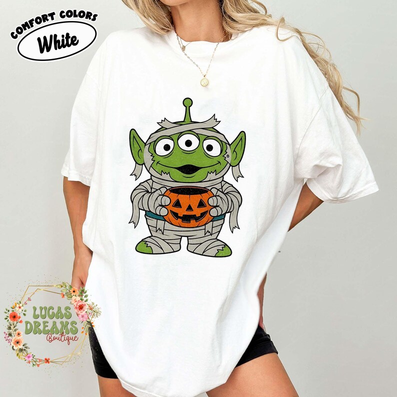 May include: White t-shirt featuring a green alien dressed as a mummy holding an orange jack-o'-lantern. The shirt has the words "COMFORT COLORS" and "White" in a circle at the top. The shirt is from "LUCAS DREAMS Boutique".