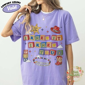 May include: A violet short-sleeve t-shirt featuring a graphic print of colorful alphabet blocks spelling "I KNEW IT I KNEW YOU". The design includes a gold sheriff badge, a red cowboy hat, a pair of cowboy boots, and a beaded bracelet with text.