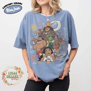 May include: A blue jean colored short-sleeve t-shirt featuring a graphic print of various animated characters including a large man with a hook, a woman, a grandmother, a small pig, and a chicken, surrounded by colorful tribal-style symbols.