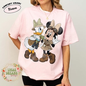 May include: Pale pink Comfort Colors t-shirt with a cartoon of Daisy Duck and Minnie Mouse in safari attire. The shirt displays the text "Comfort Colors Blossom" and "Lucas Dreams Boutique".
