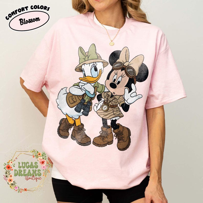 May include: Pale pink Comfort Colors t-shirt with a cartoon of Daisy Duck and Minnie Mouse in safari attire. The shirt displays the text "Comfort Colors Blossom" and "Lucas Dreams Boutique".