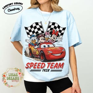 Op de afbeelding: Lichtblauw T-shirt met een afbeelding van een rode raceauto met Mickey Mouse, Donald Duck, Daisy Duck en Minnie Mouse. De woorden "SPEED TEAM 1928" staan onder de auto gedrukt, met twee geblokte vlaggen op de achtergrond.