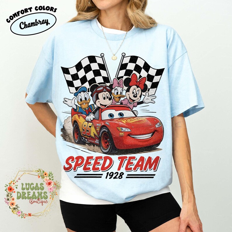 Op de afbeelding: Lichtblauw T-shirt met een afbeelding van een rode raceauto met Mickey Mouse, Donald Duck, Daisy Duck en Minnie Mouse. De woorden "SPEED TEAM 1928" staan onder de auto gedrukt, met twee geblokte vlaggen op de achtergrond.